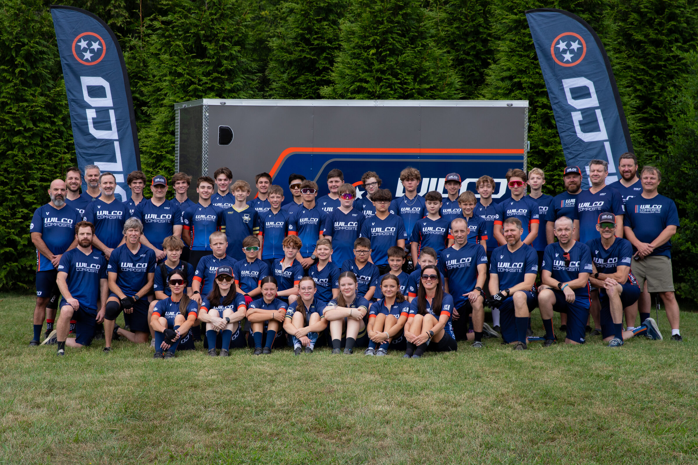 The full Wilco Composite team — riders and coaches — gathered in front of the team trailer under Wilco flags